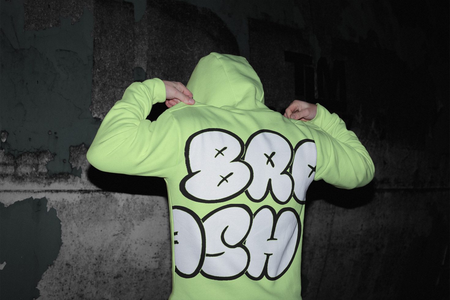 Person in neon green hoodie standing against textured wall with bold typographic design on back, ideal for mockup presentations.